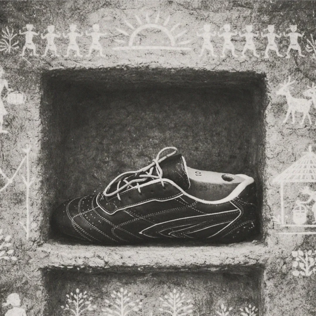 A single Sentiments sneaker placed in a wall niche, framed by traditional Indian tribal art motifs — figures, sun, and stars rendered in white pigment around the deep niche.