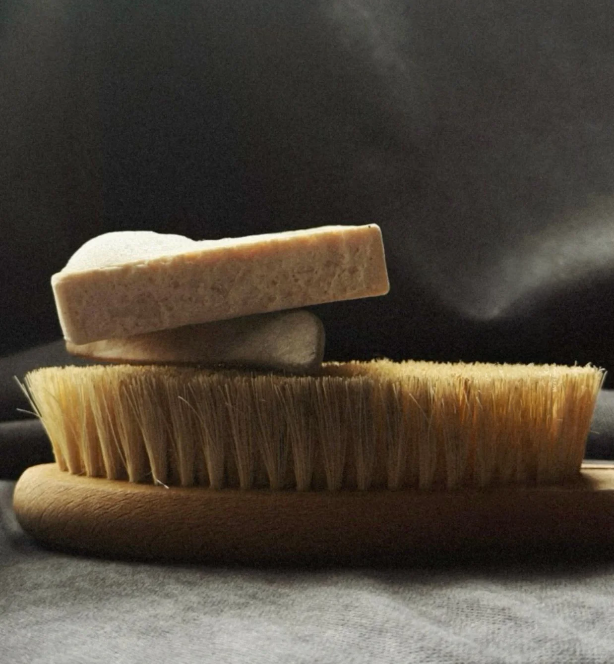 Soap bar resting on a wooden bristle brush against a dark cloth backdrop — the essentials for caring for handcrafted leather footwear.