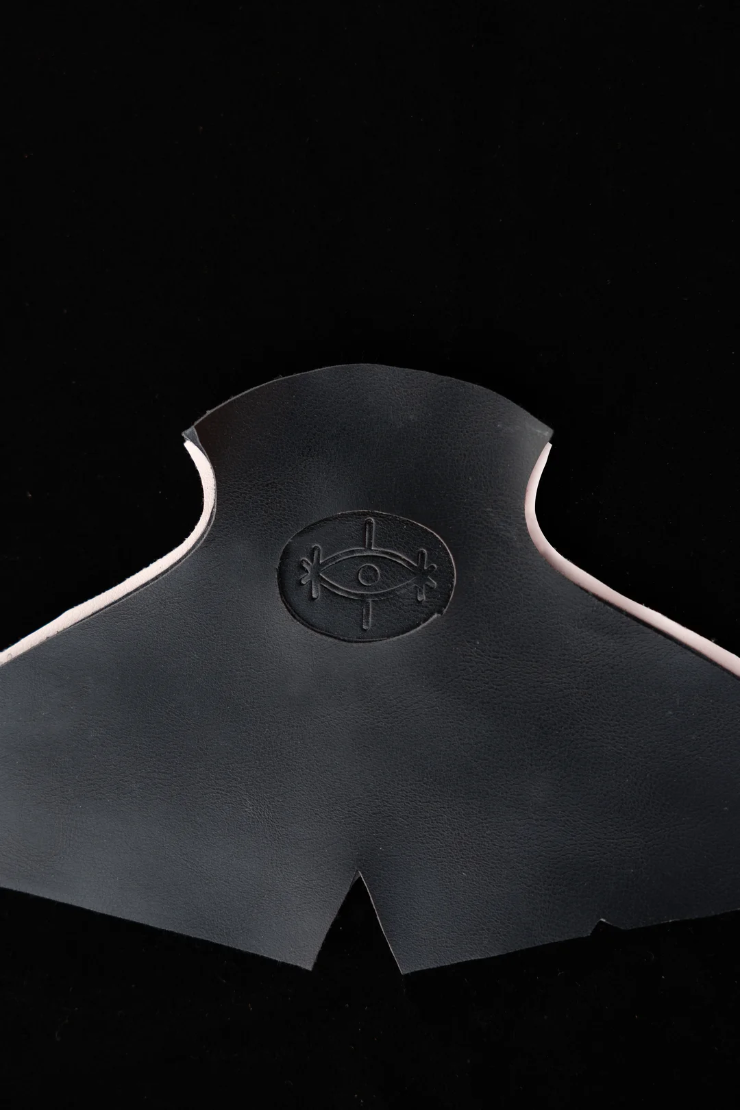A precision-cut leather component embossed with the Sentiments eye-mark logo, photographed against a deep black field — the quiet craft signature behind every pair.
