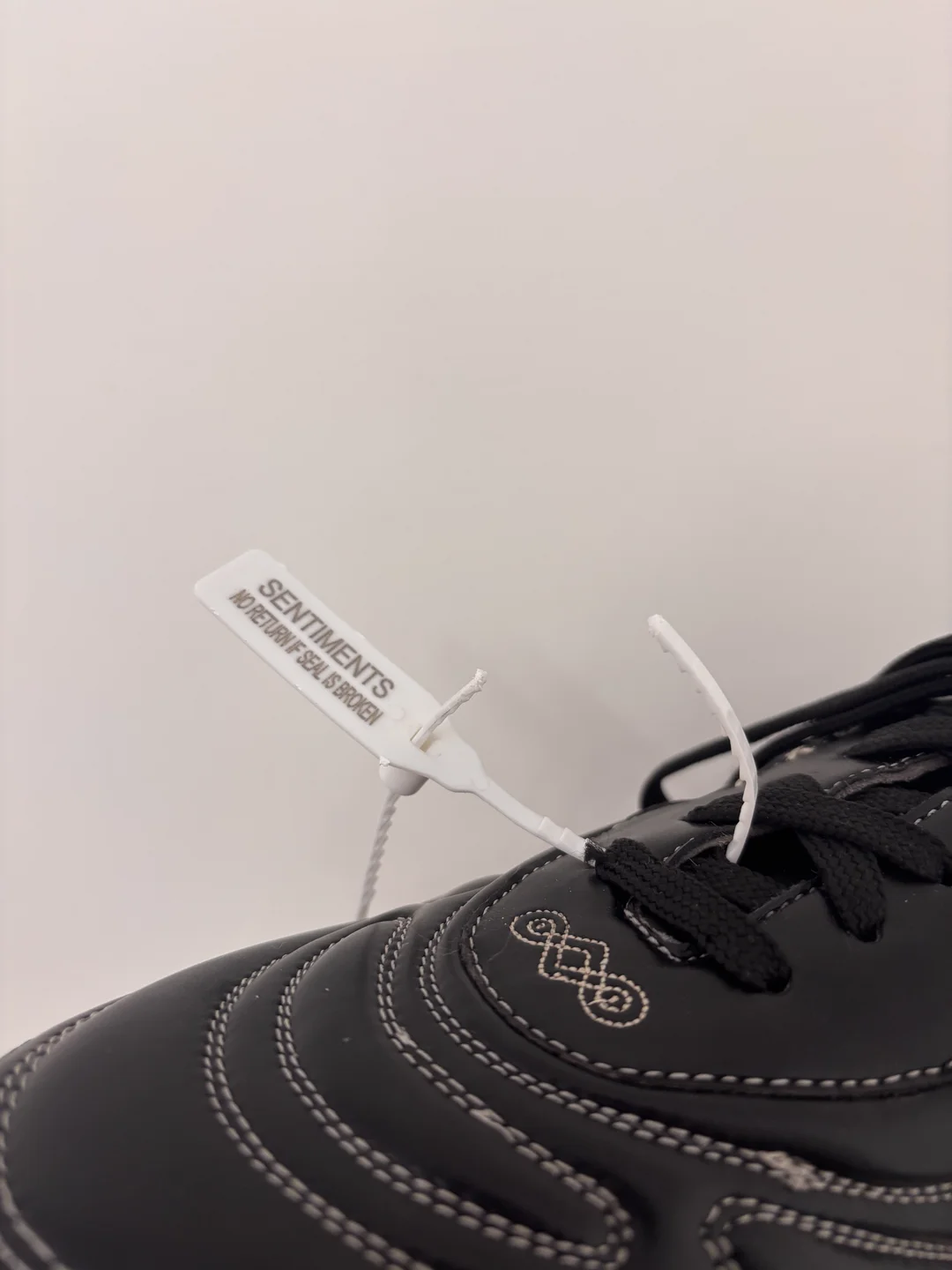 A black Sentiments shoe with the embroidered eye-mark backtab, sealed by a white tamper-evident tag reading SENTIMENTS · NO RETURN IF SEAL IS BROKEN.